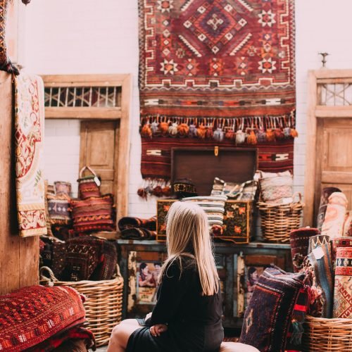 A woman sitting in a room filled with colorful Oriental carpets and woven baskets.