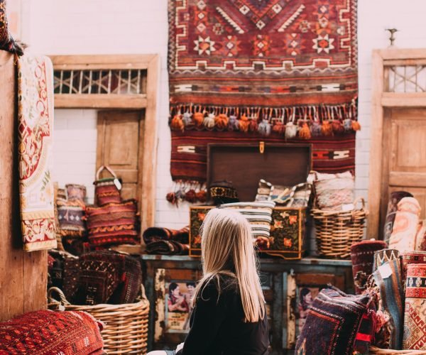 A woman sitting in a room filled with colorful Oriental carpets and woven baskets.